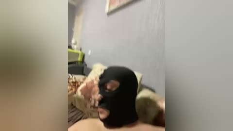 Video of a man wearing a black balaclava lying face down on a beige couch, partially visible in a dimly lit room with gray walls and a framed painting.