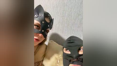 Video of a woman wearing a black leather cat mask, a beige coat, and a black balaclava, partially obscured by a blurred foreground.