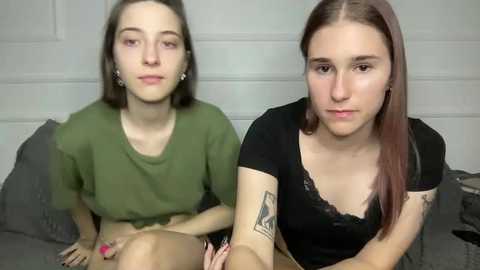 Video of two young women, one with shoulder-length brown hair in a green t-shirt, the other with straight brown hair in a black top, sitting on a bed against a white brick wall.