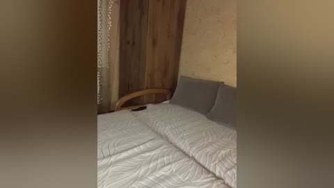 A video of a dimly lit, rustic bedroom with a wooden headboard, beige textured wallpaper, and a bed covered in a white, wavy-patterned quilt. A black smartphone lies on the bed, partially visible. The ambiance is cozy and somewhat dated.