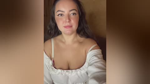 Video of a young Caucasian woman with long, dark hair, wearing a white off-shoulder top that reveals ample cleavage, set against a blurred, warm-toned background.