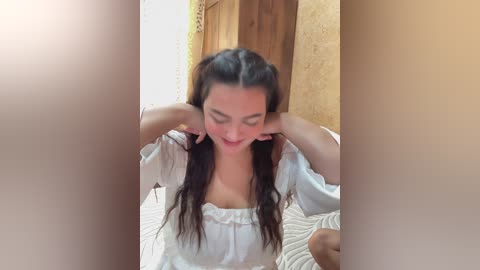 Video of a young woman with long dark hair in pigtails, wearing a white blouse, sitting on a bed with a beige quilt. Background features a wooden door and textured yellow wall.