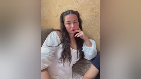 Video of a woman with long, dark hair, fair skin, and a medium build, wearing a white blouse and a textured, dark fabric. She sits on a beige cushioned chair against a textured beige wall, with a contemplative expression.