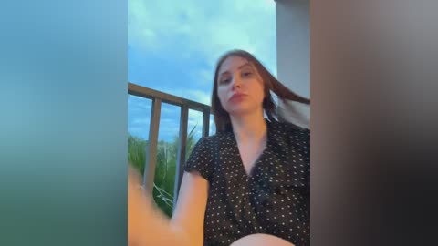 Video of a young woman with long brown hair, wearing a black polka-dot dress, standing on a balcony with a cloudy blue sky background.