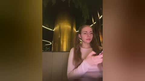 Video of a young woman with long, light brown hair in pigtails, wearing a white long-sleeved shirt, seated indoors, surrounded by warm, dim lighting and a large, shiny, golden cylindrical object.