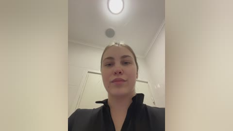 A video of a young Caucasian woman with light skin and straight, blonde hair, wearing a black robe, standing in a minimalist, well-lit bathroom with white walls and a ceiling light.