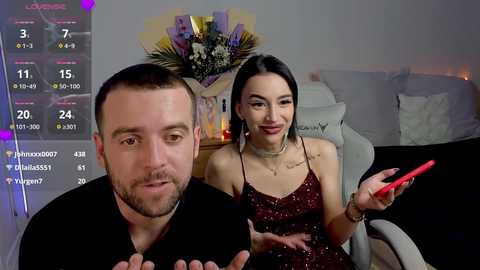 Video of a man and woman in a living room, both holding red phones, man with short hair and beard, woman with long dark hair, both smiling.