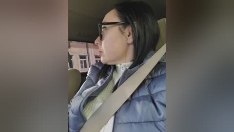 Video of a woman with long dark hair, wearing glasses and a blue puffer jacket, sitting in a car with a beige seatbelt. Background shows a window with a white frame.