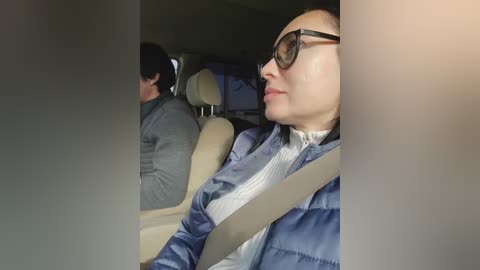 A video shows an Asian woman with glasses, wearing a blue puffer jacket and white shirt, sitting in a car, looking to the right. The interior is dimly lit with beige seats.