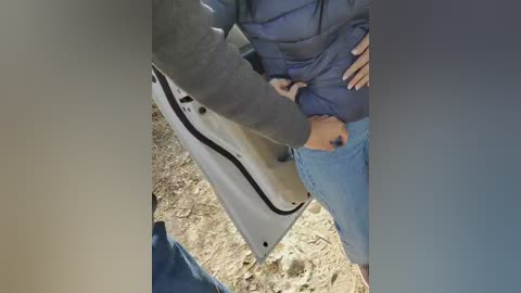 Video of a person's hands and lower body, showing a gray jacket, blue jeans, and a black gun partially visible in a waistband, standing on a gravelly ground.