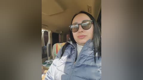 Video of a woman with straight black hair, wearing large round glasses and a blue puffer jacket, seated inside a car with a colorful, striped blanket in the background.