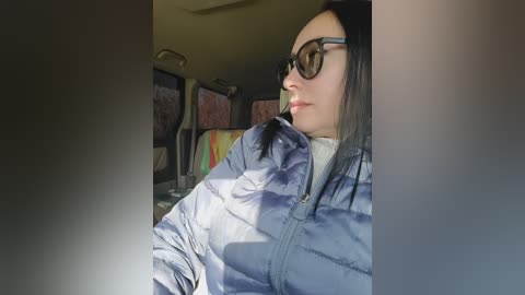 A video of a woman with long black hair, wearing large sunglasses and a puffy blue jacket, sitting in a car. The background shows a blurred, colorful, abstract painting on the wall.