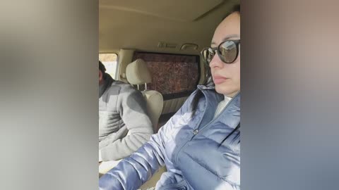 Video of a woman in sunglasses, blue jacket, and dark hair, seated in a car with beige seats and a red-patterned wall in the background.