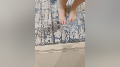 A video showing a pair of bare feet standing on a textured, abstract-patterned rug with white, blue, and grey hues. The feet are positioned centrally, and the background reveals a beige, tiled floor. The image has a soft, slightly blurred edge effect.