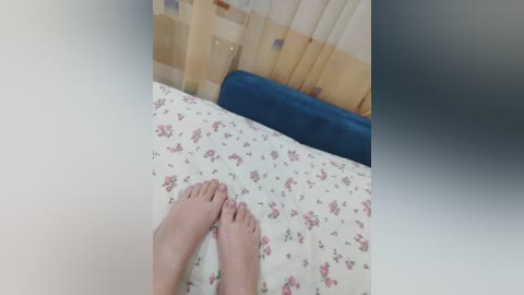 Video of a pair of bare feet on a bed with a floral-patterned white sheet. The bed features a blue cushioned headboard and beige curtains in the background.
