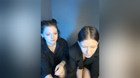 Video of two women in black, standing in a dimly lit, narrow corridor. One with dark hair styled in two buns, wearing a black jacket, looks to the side. The other, with dark hair pulled back, gazes downward, wearing a black coat.
