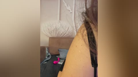 Video of a woman with shoulder-length brown hair, partially visible, standing in a dimly lit room. She's wearing a black top and jeans. A beige wall with a white, textured lampshade and a partially visible pink vibrator on a bed.