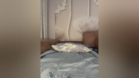 Video of a cozy, modern bedroom with a light blue quilted bedspread, a pale green pillow, and beige headboard, featuring a decorative wall design with abstract white branches and a large, beige, leaf-shaped pillow.