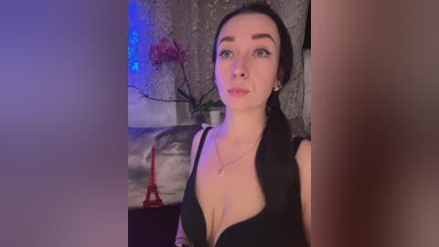 Video of a fair-skinned woman with dark hair, wearing a black dress, posing indoors. Background features a gray wall, a red Eiffel Tower, and blue fairy lights.