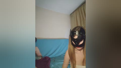A video of a woman with long brown hair wearing a black leather cat mask, standing in a room with beige walls and a blue sofa, holding a phone.