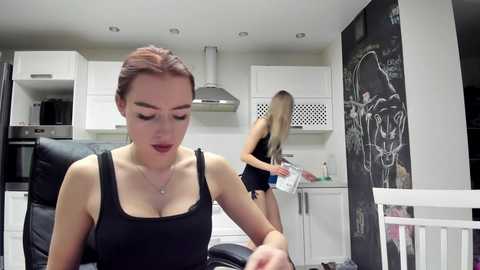 Video of a woman with auburn hair, wearing a black tank top, in a modern kitchen. Another woman, with blonde hair, is seen in the background.