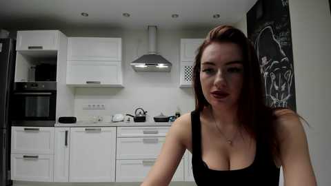 Video of a fair-skinned, red-haired woman with a slim figure, wearing a black tank top, in a modern, white kitchen with stainless steel appliances and a blackboard wall.