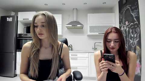 Video of two young women in a modern kitchen. One with long blonde hair, wearing a black tank top, sits on a black chair. The other, with long red hair, wears a black top and gazes at a smartphone.
