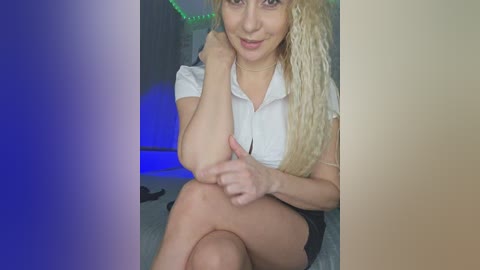 A video of a young woman with long blonde hair, fair skin, and a slender build, wearing a white shirt and black shorts, sitting on a couch with blue LED lights in the background.