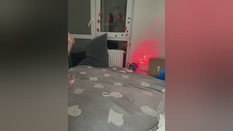 Video of a messy bedroom with a grey bedspread, scattered clothes, and a red string of fairy lights on a white radiator.