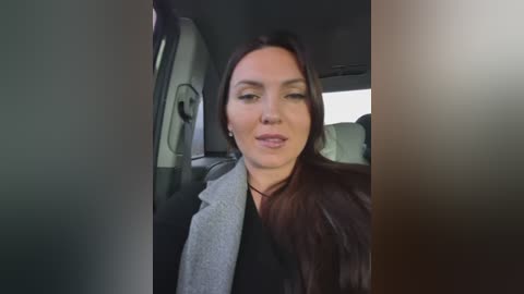A video of a light-skinned woman with long, dark hair in a car, wearing a gray scarf and black top. She has a neutral expression. The background shows the car's interior.