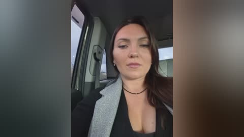 A video of a woman with long brown hair, wearing a black top and a gray blazer, sitting in a car with the window open.