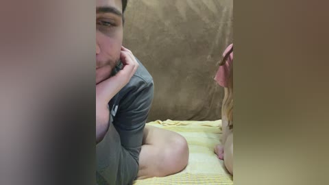 A video of a man with a beard, wearing a gray shirt and shorts, sitting on a yellow quilt. He's partially obscured by a brown curtain. A woman with long, blonde hair, wearing a pink dress, is seen from the back.