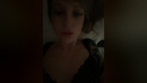 A dimly lit video of a woman with pale skin and brown hair, wearing a black lace top, captured from a slightly blurry angle, giving a mysterious, intimate atmosphere.
