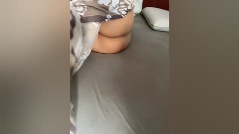 A video of a woman's lower body, wearing gray and white floral-patterned lingerie, sitting on a bed with gray sheets, a pillow, and a red wall in the background.
