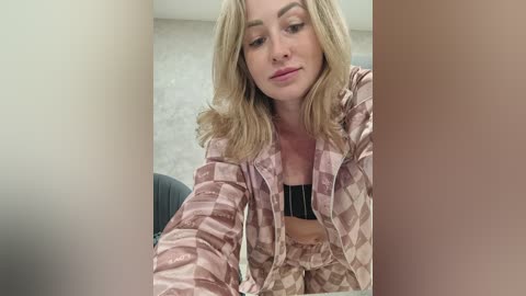 A video of a blonde woman with fair skin, wearing a pink and beige checkered pajama set, sitting on a bed, looking down.