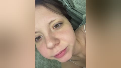 Video of a fair-skinned woman with brown hair and light makeup, slightly flushed cheeks, wearing a beige top, with a blurred background.