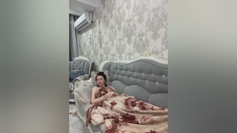 A video of a woman in a luxurious, ornate bedroom, covered in a beige bedspread with red floral patterns. She is lying on a tufted gray upholstered bed with white accents, under a gray and white damask-patterned wallpaper.