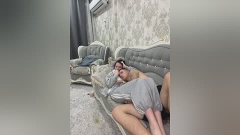 Video of a dilapidated room with two men, one lying on a worn-out couch, the other sitting beside him, wearing gray sweatpants. Wallpaper and furniture are old and dirty, contributing to a sense of neglect and poverty.