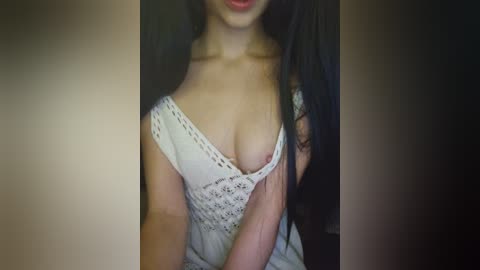 Video of a woman with long black hair, wearing a white, off-the-shoulder dress with lace, revealing cleavage, in a dimly lit room.