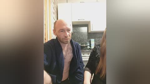 Video of a bald, bearded man in a blue robe, seated in a modern kitchen with white cabinets and dark granite countertops.