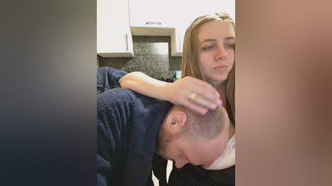 A video shows a woman with long blonde hair and a man with short hair, both in casual clothing, sharing an intimate moment in a modern kitchen with white cabinets and a dark granite backsplash.