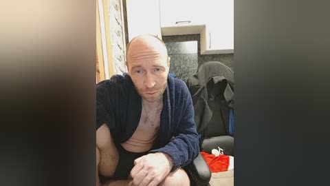 Video of a bald, bearded man in a blue robe, sitting on a bed with a grey chair and a black jacket in the background.