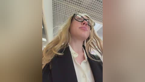 Video of a Caucasian woman with long blonde hair, wearing black-framed glasses and a black blazer over a white blouse. She stands in a tiled-ceilinged room, looking upwards with a neutral expression.