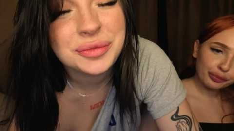 Video of two young women with pale skin, one with straight black hair and the other with red hair, both smiling. The background is dark and blurry.