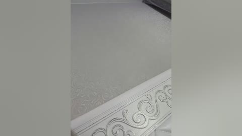 Video of a ceiling with a textured, light gray surface. A decorative, white cornice with intricate, swirling patterns runs along the top edge. The room's walls and floor are not visible.
