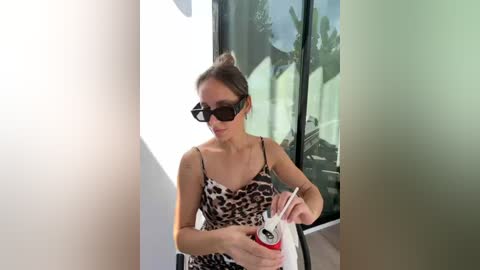 Video of a light-skinned woman with brown hair in a bun, wearing black sunglasses and a leopard-print dress, holding a white and red striped shopping bag. She stands in a bright, modern room with large windows.