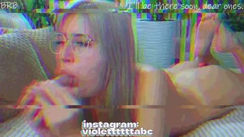 A blurry, colorful, digital video of a nude, fair-skinned woman with long hair and glasses, lying on a sofa, licking a large, erect penis. Text overlays read, \"I'll be there soon, wear ones.\