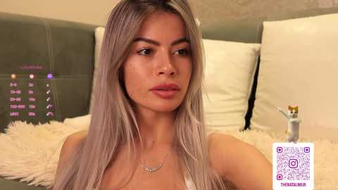 Video of a young woman with long, platinum blonde hair, fair skin, and full lips, wearing a white bra, sitting on a plush white rug in a modern bedroom.