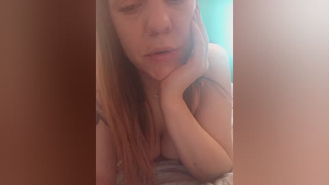 A video of a topless woman with fair skin and long red hair, resting her head on her hand, in a dimly lit room with a teal wall.