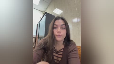 A video of a young woman with long, dark hair, fair skin, and a neutral expression, wearing a brown top, taken indoors near a wall and a TV screen.